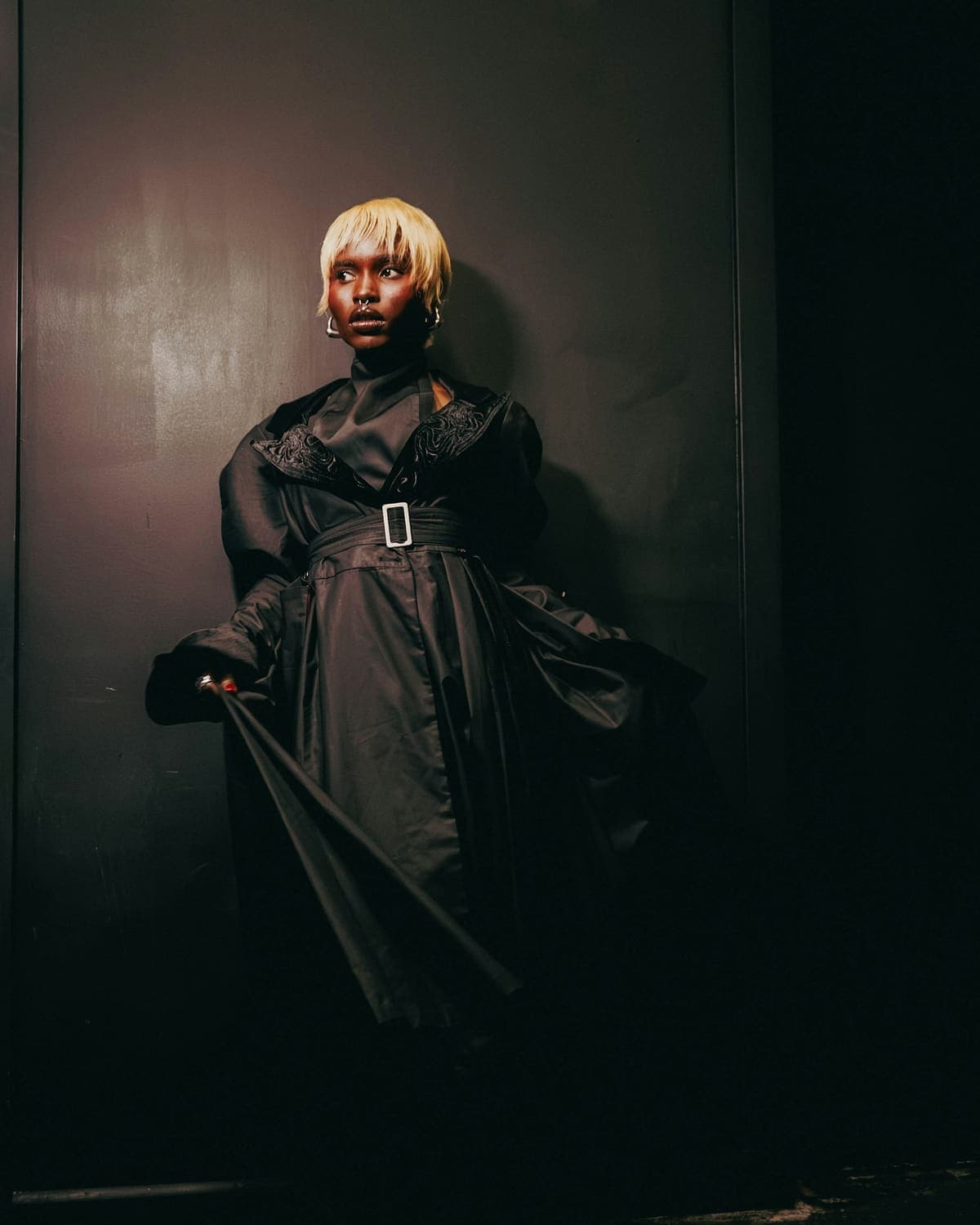 Model in a black satin coat holding the hem, in a dark room