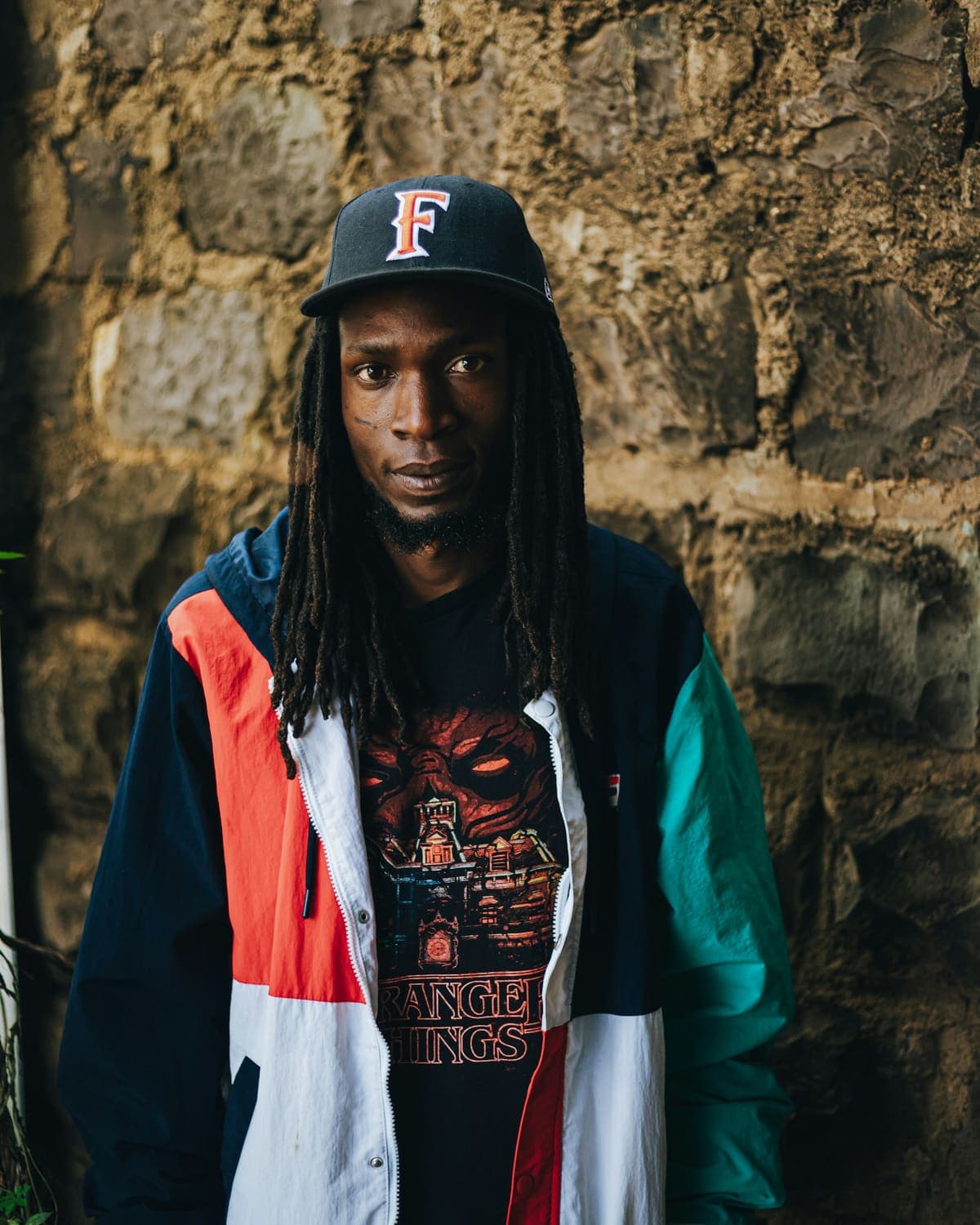 Model with locs in a colour-block jacket and cap against a stone wall